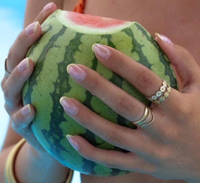 May include: A halved watermelon with green and dark green striped rind and red flesh is held by hands with long, manicured nails. The hands wear gold rings, one with a floral design. A gold bracelet is visible.