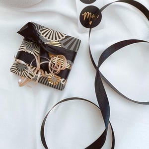 May include: A wrapped gift with black and gold patterned paper and a black bow. Gold ribbon and black ribbon are also present. Two black circular stickers with gold text that reads "Merci" are visible.