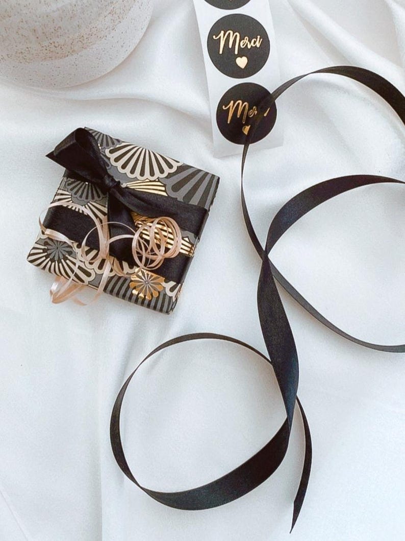 May include: A wrapped gift with black and gold patterned paper and a black bow. Gold ribbon and black ribbon are also present. Two black circular stickers with gold text that reads "Merci" are visible.