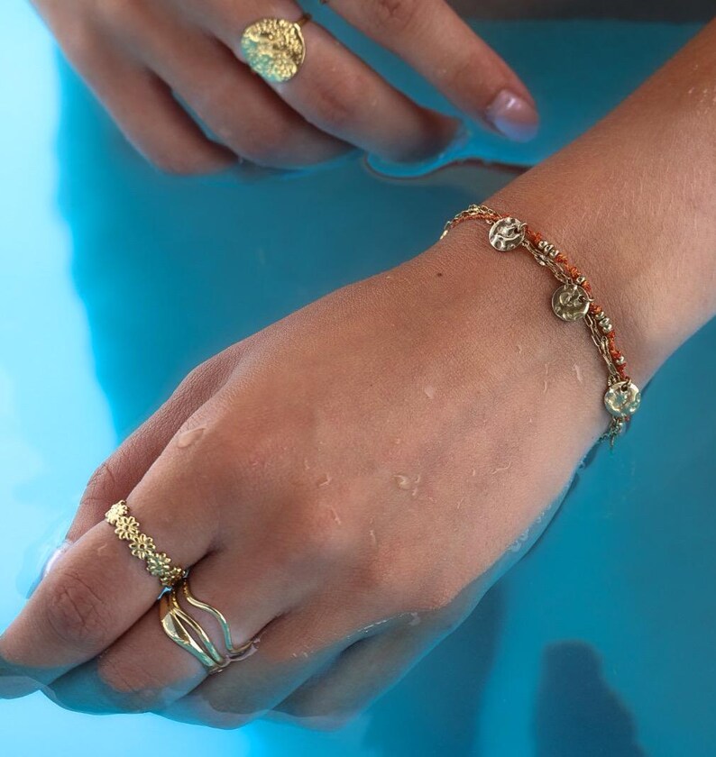 May include: A gold-toned jewelry set featuring a textured ring, a delicate ring with floral details, and a bracelet with coin-shaped charms. The jewelry is displayed against a blue background, creating a striking contrast. The pieces are designed with intricate details.