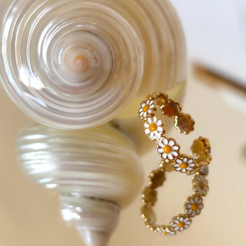 May include: A gold-toned daisy ring with white petals and yellow centers. The ring is open and adjustable. A large, pearlescent seashell is in the background, reflecting the ring's image.