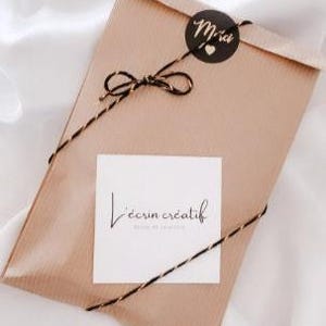 May include: A wrapped package in a brown paper envelope, tied with black and gold twine. A small black sticker with the word "Merci" and a heart is on the top. A white square with the text "L'&eacute;crin cr&eacute;atif" is on the front.
