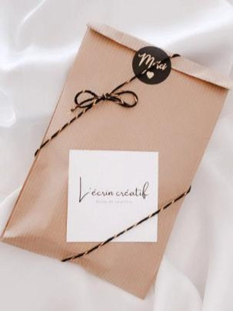 May include: A wrapped package in a brown paper envelope, tied with black and gold twine. A small black sticker with the word "Merci" and a heart is on the top. A white square with the text "L'&eacute;crin cr&eacute;atif" is on the front.