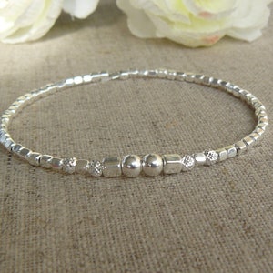 Könnte beinhalten: Ein silbernes Armband mit kleinen quadratischen und runden Perlen. Das Armband hat ein Stretch-Design und verfügt über einige größere runde Perlen und funkelnde Akzente. Der Schmuck wird auf einer neutralen Leinenoberfläche präsentiert.