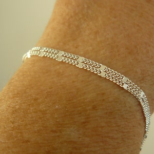 May include: A delicate silver bracelet with two parallel strands of small, rectangular links. The bracelet is worn on a person's wrist, showcasing its simple yet elegant design. The bracelet's length is approximately 7 inches.