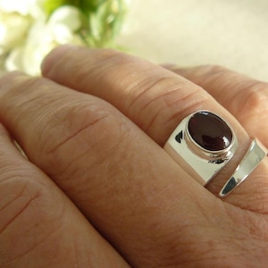 Puede incluir: Un anillo de plata con una piedra ovalada de color rojo oscuro engastada en un engaste de bisel. El anillo tiene un diseño único con una banda que se envuelve alrededor del dedo.