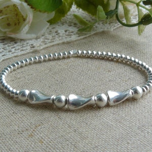 Puede incluir: Un brazalete de plata con pequeñas cuentas redondas y cuentas de plata más grandes y curvadas. El brazalete está sobre un fondo de tela marrón claro.
