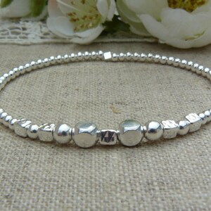 May include: A silver bracelet featuring small round beads and larger square beads. The bracelet is made of metal and has a simple design.
