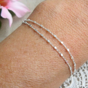 Peut inclure: Un bracelet argenté délicat avec deux rangées de petites perles réfléchissantes espacées régulièrement. Le bracelet est porté sur un poignet. Une fleur rose tendre et des feuilles vertes sont en arrière-plan.