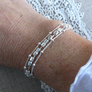 Puede incluir: Un brazalete de plata con tres cadenas y pequeñas cuentas cuadradas.