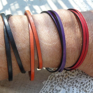 May include: A collection of thin leather bracelets in various colors, including black, orange, purple, and red. The bracelets are wrapped around a wrist, showcasing their simple, minimalist design. The bracelets are a stylish accessory.