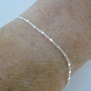 Peut inclure: Un bracelet en argent avec une chaîne fine et un design à maillons. Le bracelet est présenté sur le poignet d'une personne.