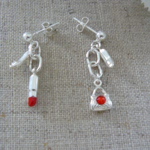 May include: A pair of silver-toned drop earrings featuring charms. One earring displays a high-heeled shoe and a lipstick tube with a red tip. The other earring features a chain link, a small cylinder, and a handbag charm with a red circular stone.