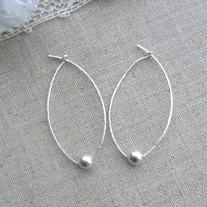May include: A pair of silver oval hoop earrings. Each earring has a small, round bead at the bottom. The earrings are presented on a neutral, textured surface.