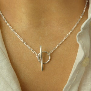 Peut inclure: Un collier en chaîne argent avec un fermoir à glissière. Le fermoir est un cercle avec une barre qui coulisse à travers lui.