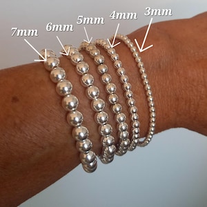 Op de afbeelding: Een close-up van een pols met zes zilveren kralen armbanden. De armbanden zijn op elkaar gestapeld, met de grootste armband onderaan en de kleinste armband bovenaan. De armbanden zijn gelabeld met hun kralenmaat in millimeters: 7 mm, 6 mm, 5 mm, 4 mm, 3 mm.