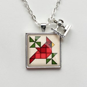May include: A silver necklace with a square pendant featuring a red cardinal bird made of geometric shapes. The pendant is surrounded by a silver frame. A small silver sewing machine charm hangs from the necklace.