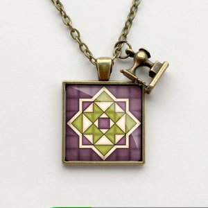 May include: A bronze necklace with a square pendant featuring a geometric quilt block design in shades of purple, green, and white. A small bronze sewing machine charm hangs from the necklace.