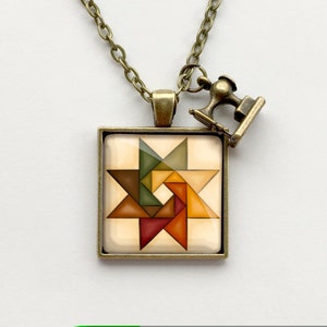 May include: A gold necklace with a square pendant featuring a colourful geometric star design. The pendant is surrounded by a bronze frame. A small sewing machine charm hangs from the necklace.