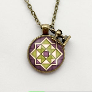 May include: A bronze necklace with a round pendant featuring a geometric design of green and purple triangles. The pendant is attached to a small bronze charm shaped like a sewing machine.