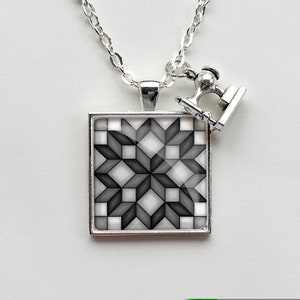 May include: A silver necklace with a square pendant featuring a black and white geometric design. A small silver charm of a sewing machine hangs from the necklace.