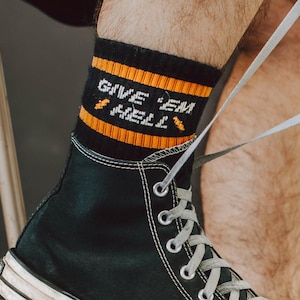 May include: Black high-top sneakers with white soles and laces. The sneakers are paired with black socks featuring orange stripes and the text "GIVE 'EM HELL". The classic Converse Chuck Taylor All Star design is visible.