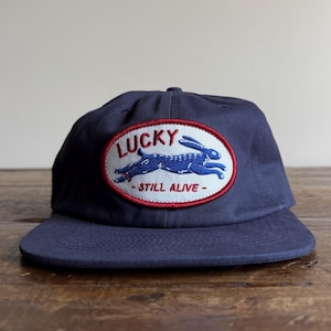 May include: Navy blue baseball cap with a curved brim. The cap features an embroidered patch with the word "LUCKY" above a blue rabbit graphic, and the words "STILL ALIVE" below. The patch is framed by a red border.