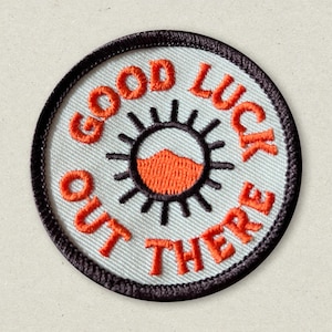 May include: A white embroidered patch with a black outline. The patch features a stylized sun with an orange center and the text "Good Luck Out There" in orange.