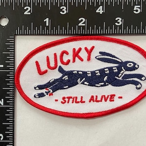 May include: Oval patch with a red border and white background. The patch features a navy blue rabbit leaping across the center. The word "LUCKY" is embroidered above the rabbit, and "- STILL ALIVE -" below.