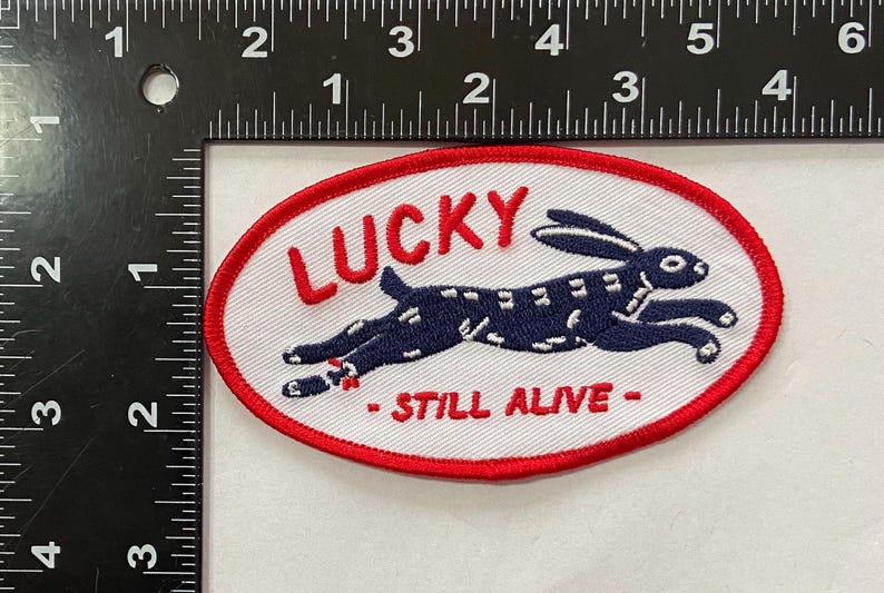 May include: Oval patch with a red border and white background. The patch features a navy blue rabbit leaping across the center. The word "LUCKY" is embroidered above the rabbit, and "- STILL ALIVE -" below.
