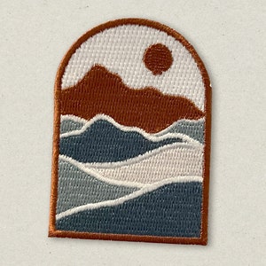 May include: An embroidered patch with a white background, featuring a brown mountain range with a red sun, and a blue and gray layered wave pattern.