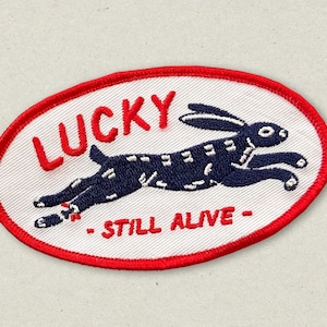 May include: Oval patch with a white background and a red border. The patch features a navy blue rabbit leaping across the center. The word "LUCKY" is in red above the rabbit, and "- STILL ALIVE -" is below.