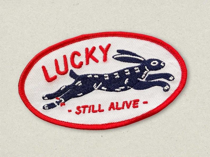 May include: Oval patch with a white background and a red border. The patch features a navy blue rabbit leaping across the center. The word "LUCKY" is in red above the rabbit, and "- STILL ALIVE -" is below.