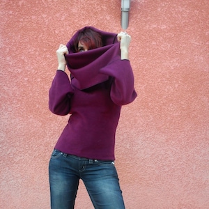 May include: A deep purple hooded sweater with long sleeves. The sweater is paired with blue denim jeans. The person is holding the hood up, partially obscuring their face. The background is a textured, salmon-colored wall.