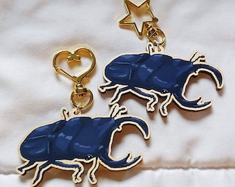 May include: Two wooden keychains featuring a dark blue stag beetle design. Each keychain has a gold-colored metal loop and a heart-shaped or star-shaped charm.