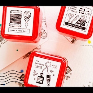 Peut inclure: Trois tampons en caoutchouc carrés rouges avec des illustrations et du texte en noir et blanc. Les tampons présentent les phrases "Enjoy Handmade Have a Nice Day!"  "Hello Small Things! How Are You?" et "My Favorite Things Have Fun!"