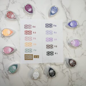 May include: A collection of ten Versa Magic ink pads in various colors, including purple, pink, red, brown, and green. Each pad has a teardrop shape and features a patterned design on the front. The ink pads are arranged in two rows on a white marble surface.