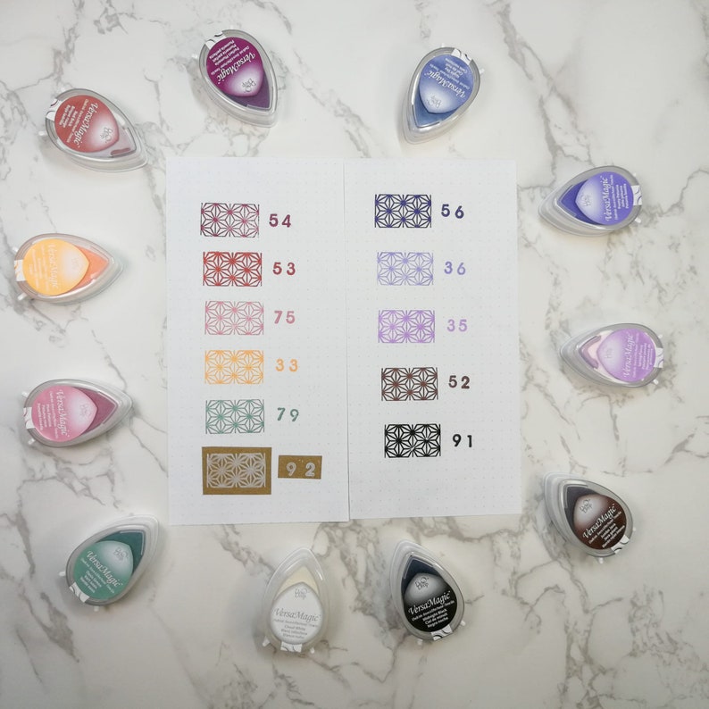 May include: A collection of ten Versa Magic ink pads in various colors, including purple, pink, red, brown, and green. Each pad has a teardrop shape and features a patterned design on the front. The ink pads are arranged in two rows on a white marble surface.
