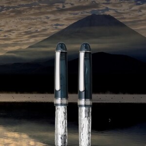 May include: Two silver and blue fountain pens with a textured pattern. The pens are against a backdrop of a mountain and a cloudy sky.