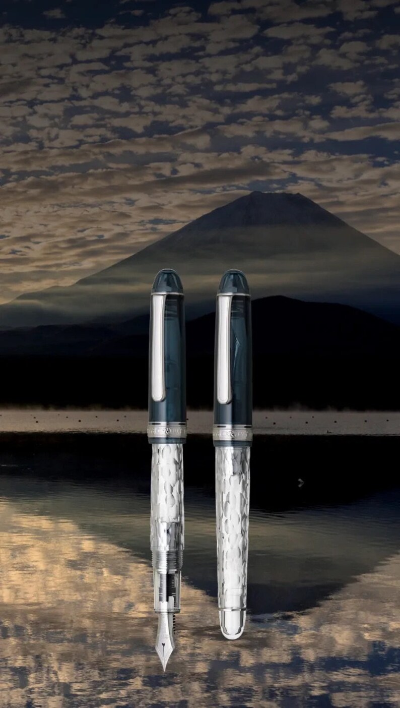 May include: Two silver and blue fountain pens with a textured pattern. The pens are against a backdrop of a mountain and a cloudy sky.