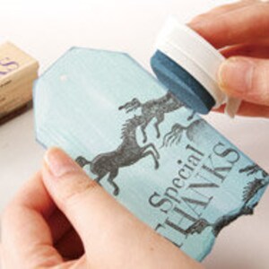 May include: A blue and white rectangular stamp with a design of two horses running. The stamp is being used to create an impression on a blue and white tag with the text "Special Thanks".