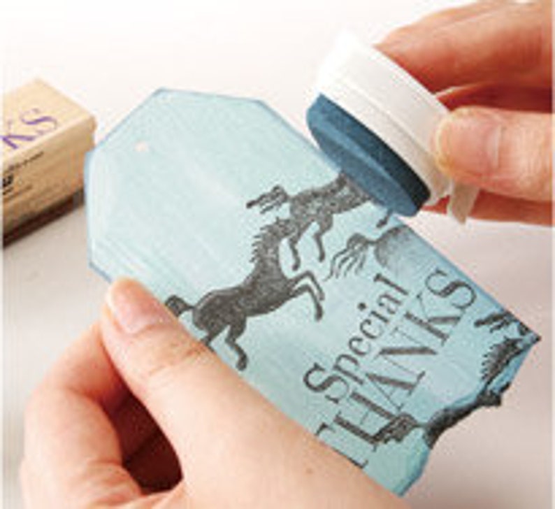 May include: A blue and white rectangular stamp with a design of two horses running. The stamp is being used to create an impression on a blue and white tag with the text "Special Thanks".