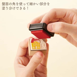 Puede incluir: Un sello de madera con un dise&ntilde;o de casa amarilla y roja. Se est&aacute; utilizando una almohadilla de tinta roja separada para estampar el dise&ntilde;o.