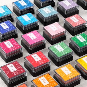 Puede incluir: Surtido de almohadillas de tinta de colores con texto e ilustraciones japonesas. Las almohadillas de tinta son de varios colores, incluyendo rojo, rosa, azul, verde, amarillo y naranja. Las almohadillas de tinta est&aacute;n en un recipiente de pl&aacute;stico transparente con una base negra.