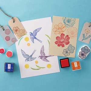 Puede incluir: Un conjunto de sellos de goma de colores con dise&ntilde;os florales y de p&aacute;jaros. Los sellos est&aacute;n dispuestos sobre una superficie azul claro con almohadillas de tinta y tarjetas en blanco.