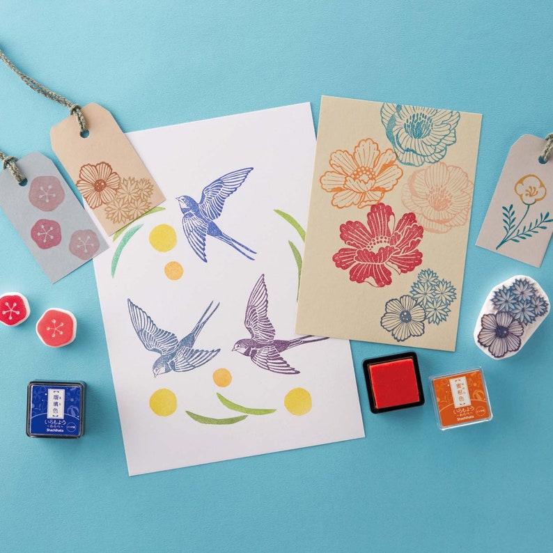 Puede incluir: Un conjunto de sellos de goma de colores con dise&ntilde;os florales y de p&aacute;jaros. Los sellos est&aacute;n dispuestos sobre una superficie azul claro con almohadillas de tinta y tarjetas en blanco.