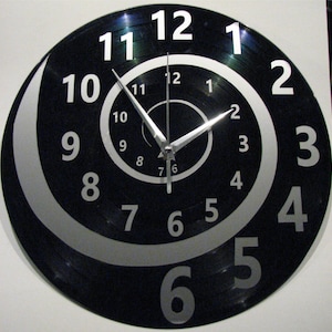 Könnte beinhalten: Eine schwarze Schallplattenuhr mit silbernen Zahlen und Zeigern. Die Zahlen sind spiralförmig um das Zifferblatt angeordnet.