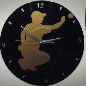 Peut inclure: Une horloge en vinyle noir avec une silhouette dorée d'une personne lançant une balle. Le cadran de l'horloge a des aiguilles argentées et des chiffres blancs.