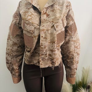 May include: A cropped camouflage jacket with a distressed hem and embellished accents on the shoulders and cuffs. The jacket features a button-up front and two chest pockets. The color palette includes shades of tan, brown, and beige.