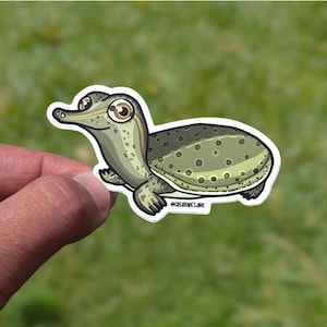 May include: A die-cut sticker featuring a cartoon illustration of a softshell turtle with a long, pointed snout and large brown eyes. The turtle has a light green body with dark green spots on its shell and the text @CREATURES.INK printed on it.